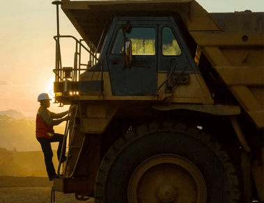 Photo of a man climbing the ladder of a truck with a huge wheel. The man is wearing Vale uniform, a green button-down shirt, an orange vest with fluorescent illumination, and a helmet.