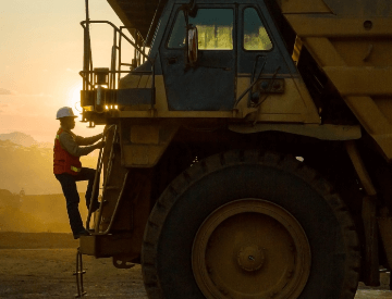 Photo of a man climbing the ladder of a truck with a huge wheel. The man is wearing Vale uniform, a green button-down shirt, an orange vest with fluorescent illumination, and a helmet.