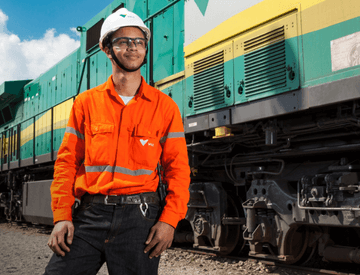 A man standing next to a green, yellow and gray train. He is wearing orange uniform with Vale logo, goggles, and a white helmet also with the company logo.