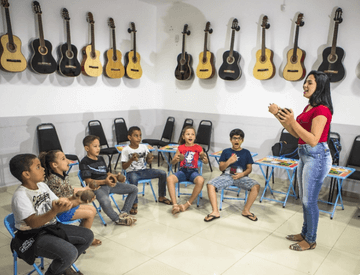 Music room with several guitars hanging on the wall. In the center, there are several children sitting in circles paying attention to a gesticulating woman.