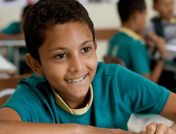 A boy in a classroom paints a sheet using crayons. He wears braces on his teeth and green and yellow uniform.