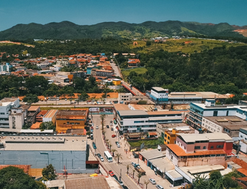 Imagem aérea de uma cidade. Em primeiro plano há uma avenida, casas e comércios e, ao fundo, é possível ver bastante vegetação e áreas montanhosas.