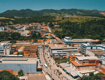 Aerial image of a city. In the foreground, there is an avenue, houses and shops, and in the background, it is possible to see a lot of vegetation and mountainous areas.