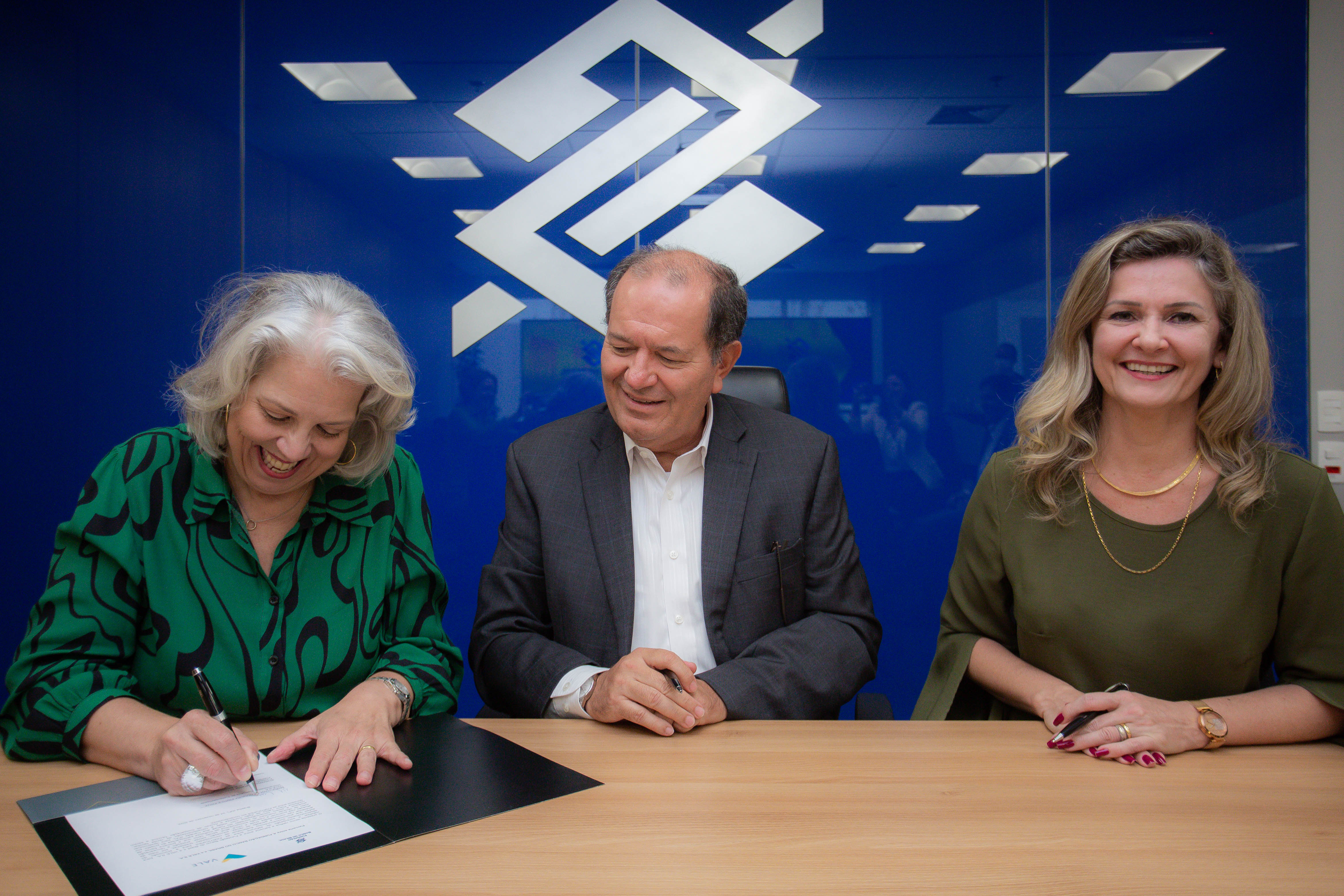 Foto de duas mulheres nas pontas e um homem no meio, sentados e apoiados em uma mesa sorrindo. A mulher da esquerda está assinando um papel.